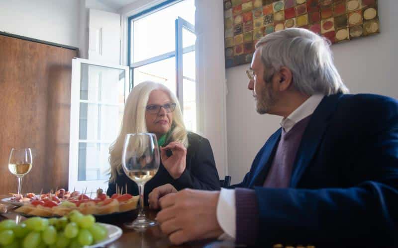 Senior couple sitting at a dining table in a bright, cozy room, enjoying appetizers and white wine while having a warm conversation.